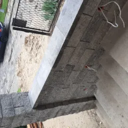 Szare kamienne płytki elewacyjne ułożone na zewnątrz budynku, widoczne wystające kable elektryczne, w tle piasek, brama i zaparkowany niebieski samochód.