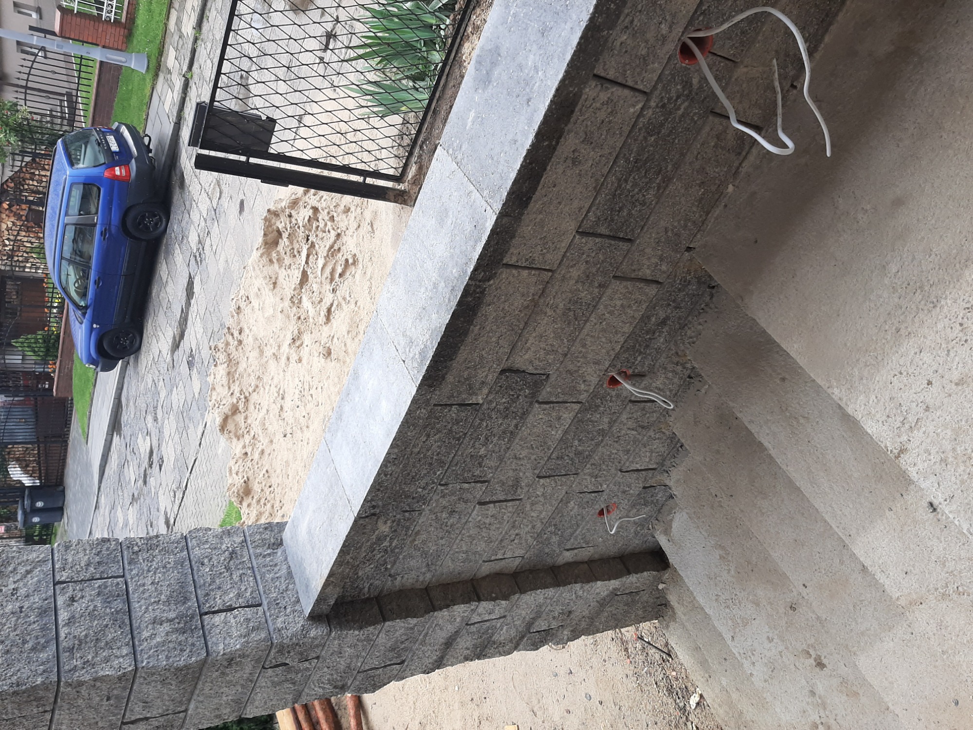 Szare kamienne płytki elewacyjne ułożone na zewnątrz budynku, widoczne wystające kable elektryczne, w tle piasek, brama i zaparkowany niebieski samochód.