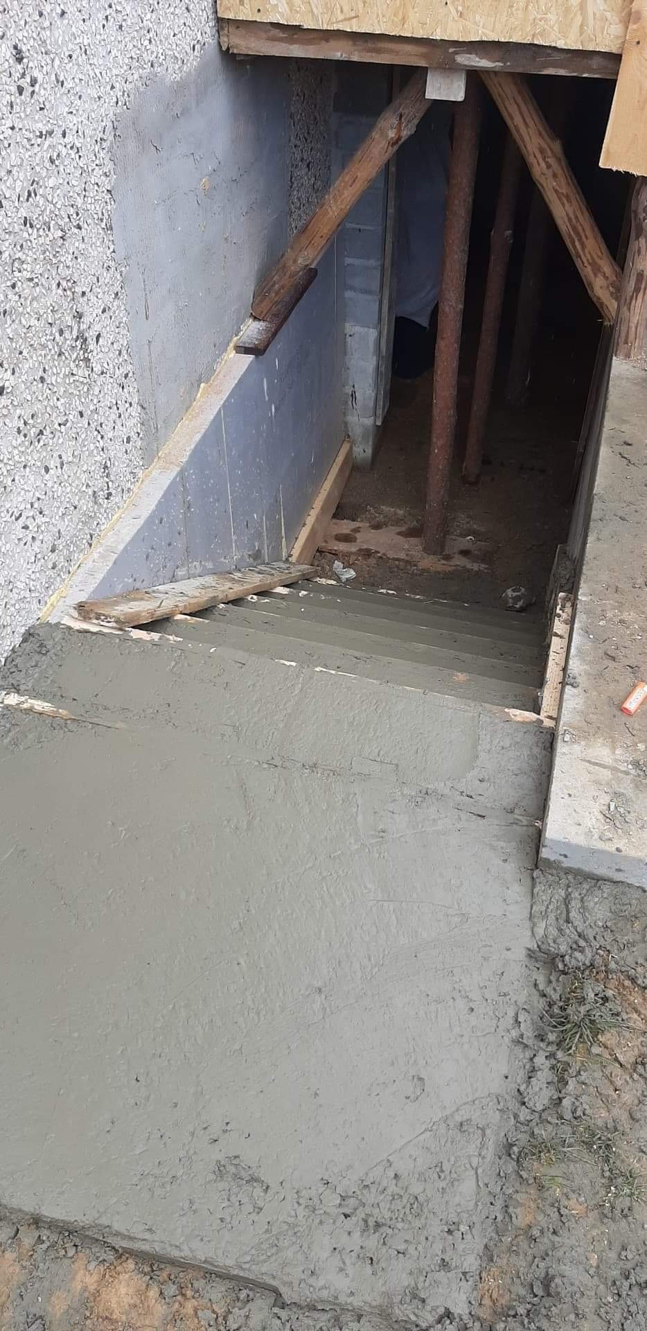 Świeżo wylane betonowe schody prowadzące do piwnicy, widoczne drewniane podpory i szalunki, surowy stan wykończenia.