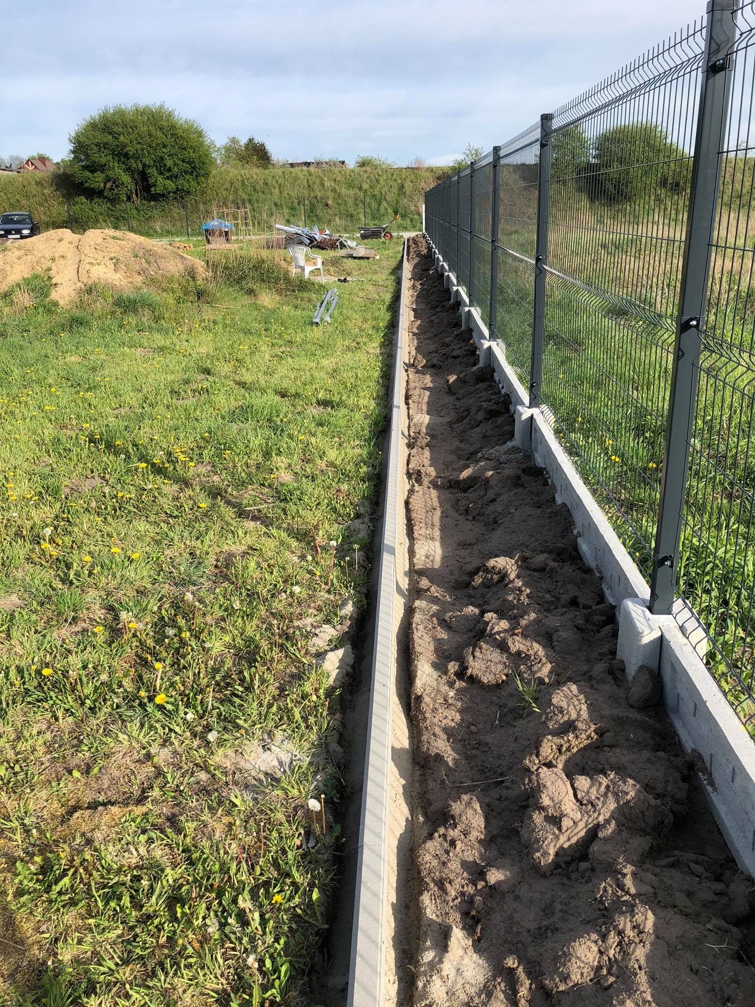 Wykop wzdłuż ogrodzenia panelowego z betonową podmurówką, przygotowany pod instalację. W tle widoczne prace ziemne i sprzęt budowlany.