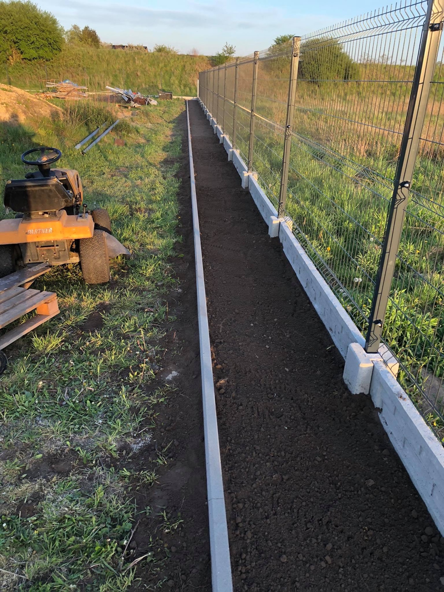Przygotowany pas ziemi obramowany betonowymi obrzeżami wzdłuż metalowej siatki ogrodzeniowej, traktor-kosiarka na trawie w tle.