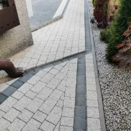 Jasnoszara kostka brukowa ułożona w geometryczny wz&oacute;r z ciemnoszarymi obrzeżami, brązowa rynna spustowa przy ścianie budynku i ozdobny żwir przy rabacie z roślinami.