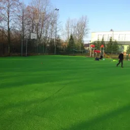 Inspektor nadzoru na boisku ze sztuczną trawą, oceniający jakość wykonania, z widoczną infrastrukturą sportową i placem zabaw w tle.