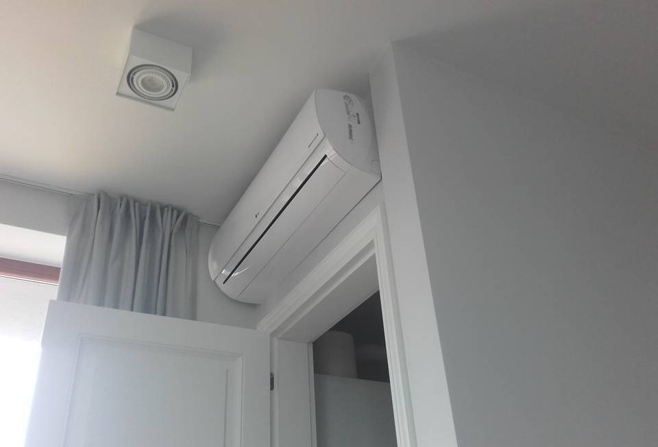 Biały klimatyzator LG zamontowany nad futryną drzwiową w jasnym pomieszczeniu, z widoczną lampą sufitową i zasłoną w tle.