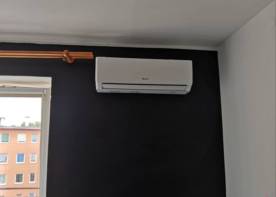 Biała jednostka klimatyzacyjna marki Gree zamontowana na czarnej ścianie w pokoju, widoczny karnisz w kolorze miedzianym i okno z widokiem na budynek.
