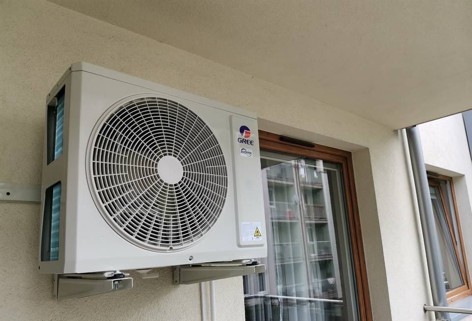 Zewnętrzna jednostka klimatyzacji marki GREE zamontowana na balkonie, widoczne uchwyty montażowe i okno z odbiciem budynku.