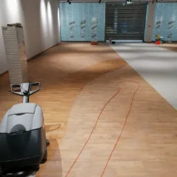 Automat szorująco-zbierający na drewnianej podłodze w przestronnym, jasnym pomieszczeniu z widoczną częścią umytą i nieumytą, kabel zasilający rozciągnięty na podłodze.