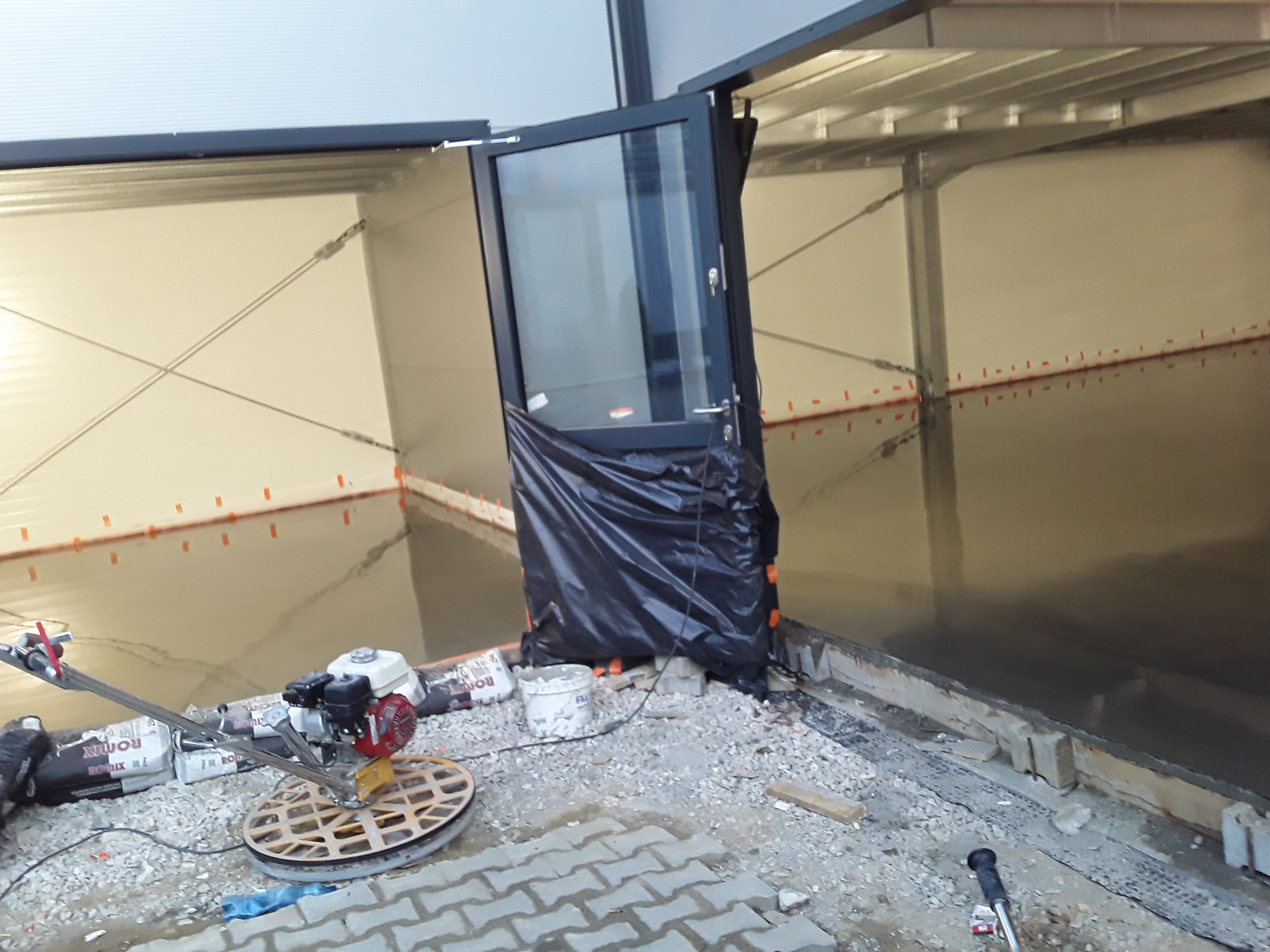 Świeżo wylana, mokra posadzka w hali, widoczna zacieraczka do betonu, worki z zaprawą i wiaderka. Drzwi zabezpieczone folią.