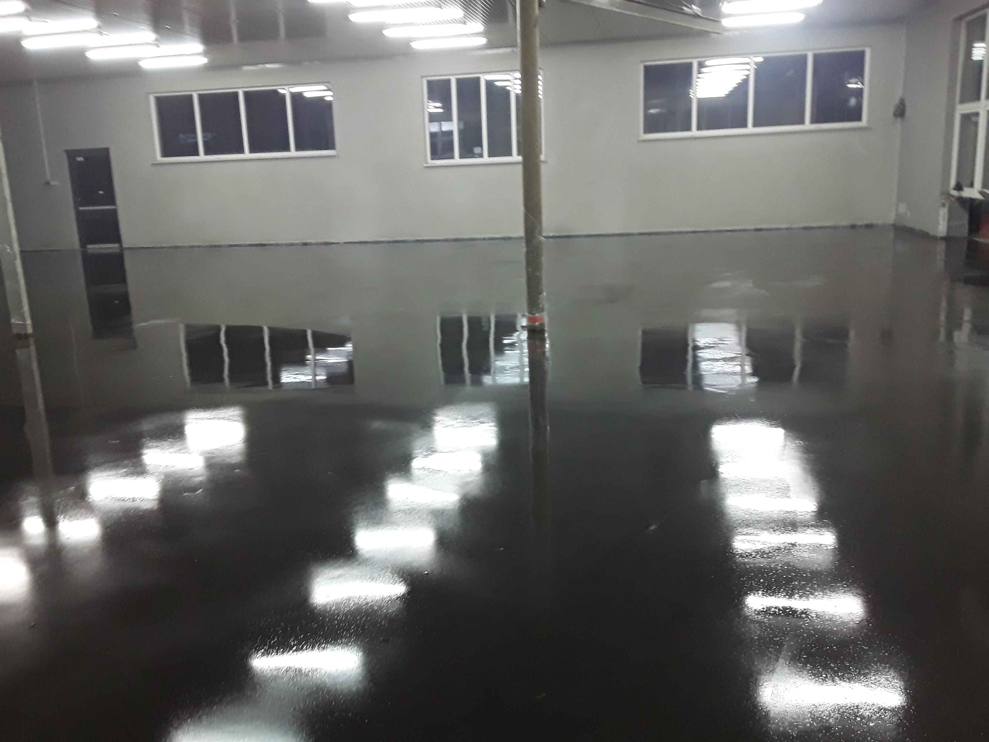 Błyszcząca, ciemna posadzka przemysłowa w hali z widocznymi oknami i oświetleniem sufitowym, odbijającym się w powierzchni podłogi.