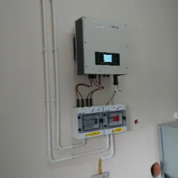 Biały inwerter solarny z wyświetlaczem i dwoma rozdzielnicami pod spodem, połączone przewodami i rurkami PCV na beżowej ścianie, obok bojler.