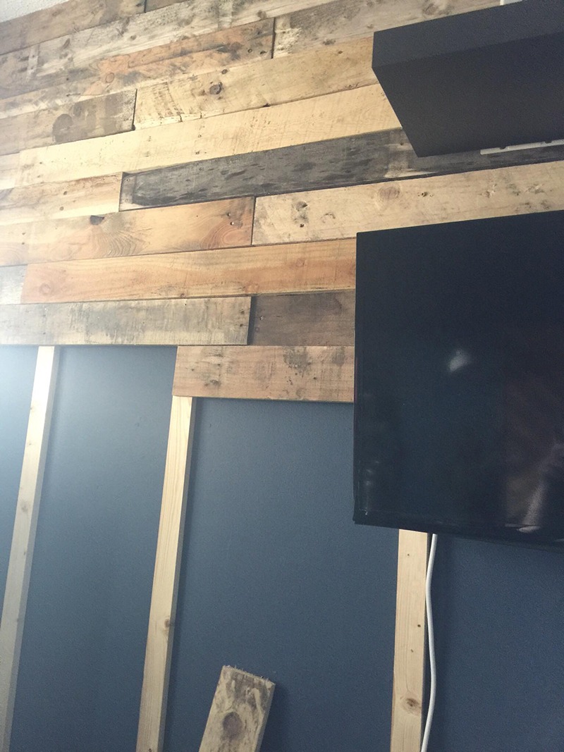 Fragment ściany z drewnianych desek o różnej kolorystyce, z widoczną konstrukcją wsporczą z surowego drewna i fragmentem telewizora, na tle pomalowanej na ciemny niebieski kolor ściany.