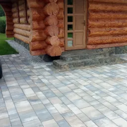 Fragment domu z bali z wejściem wyłożonym kostką brukową w odcieniach szarości i beżu, kamienne schody wejściowe.