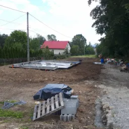 Wylany fundament z hydroizolacją i wystającymi rurami kanalizacyjnymi, widok na plac budowy z paletą i bloczkami fundamentowymi w pierwszym planie.