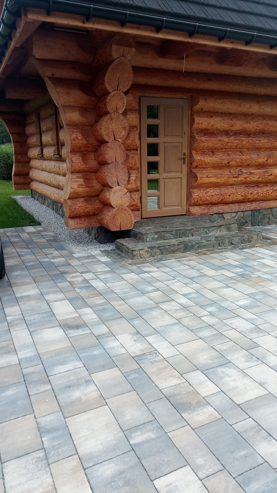 Fragment domu z bali z wejściem wyłożonym kostką brukową w odcieniach szarości i beżu, kamienne schody wejściowe.