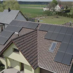 Widok z góry na dom z brązową dachówką i panelami słonecznymi, w tle inne domy i pola.