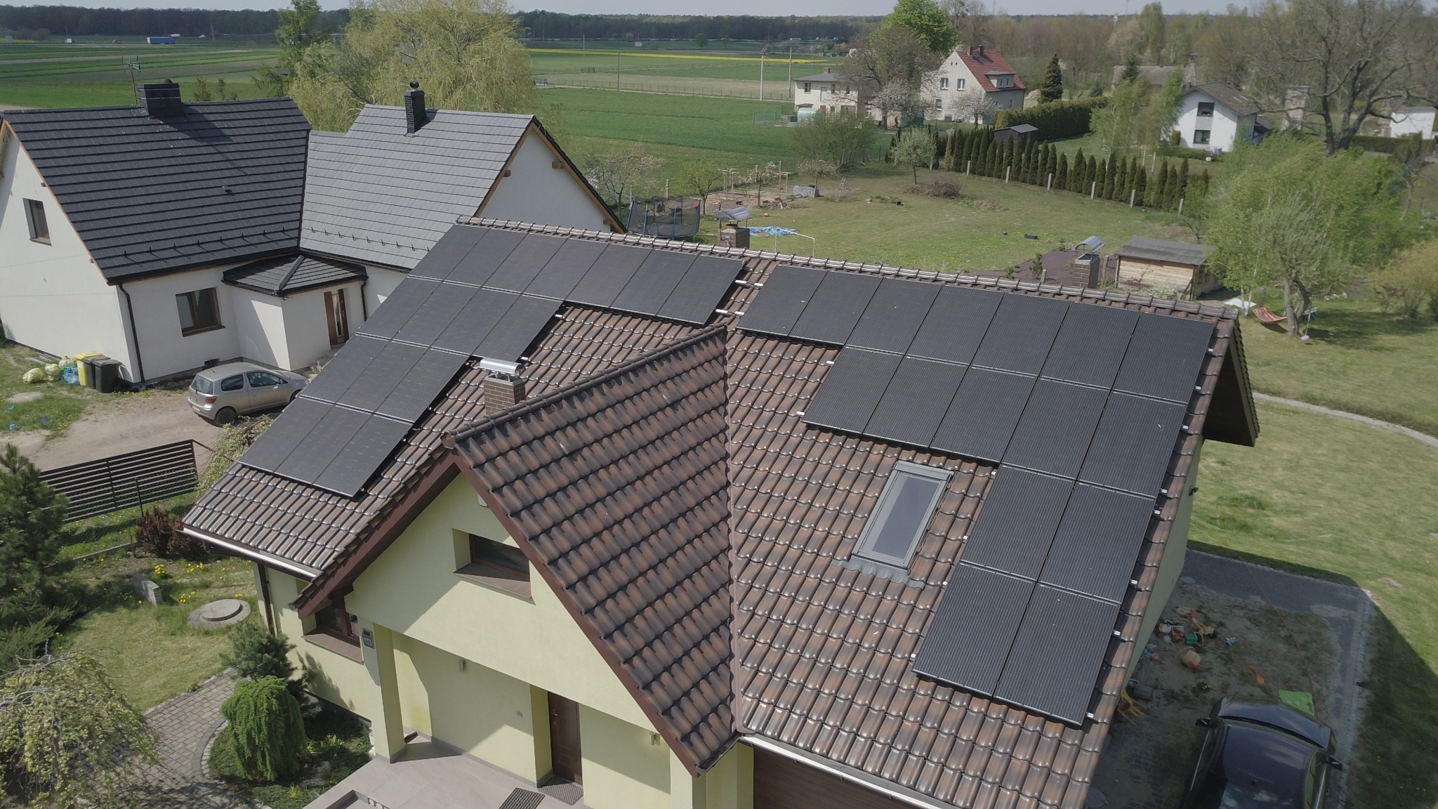 Widok z góry na dom z brązową dachówką i panelami słonecznymi, w tle inne domy i pola.
