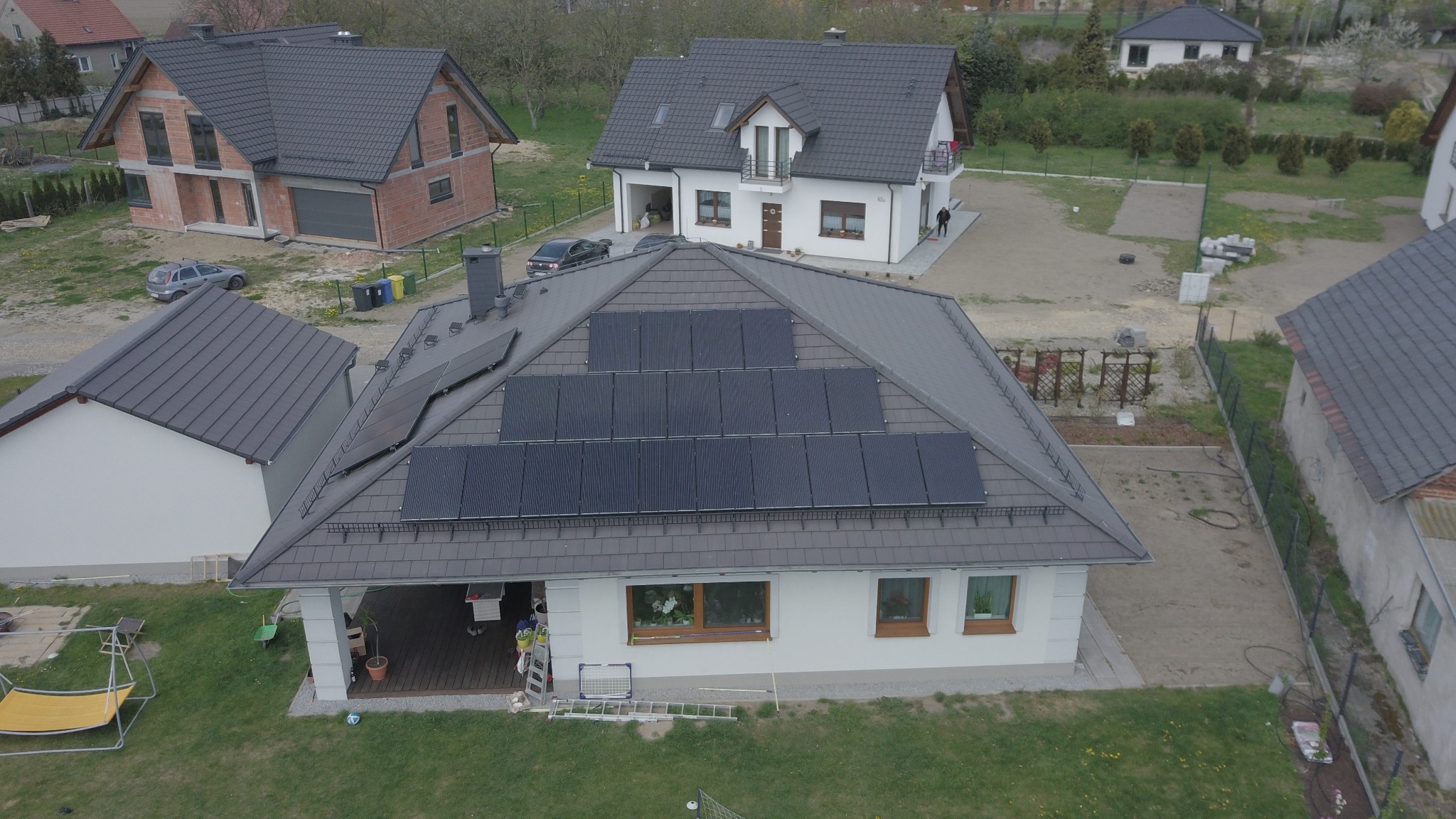 Widok z góry na dom jednorodzinny z szarym dachem pokrytym panelami słonecznymi; w tle inne domy w różnym stanie budowy i zagospodarowania terenu.