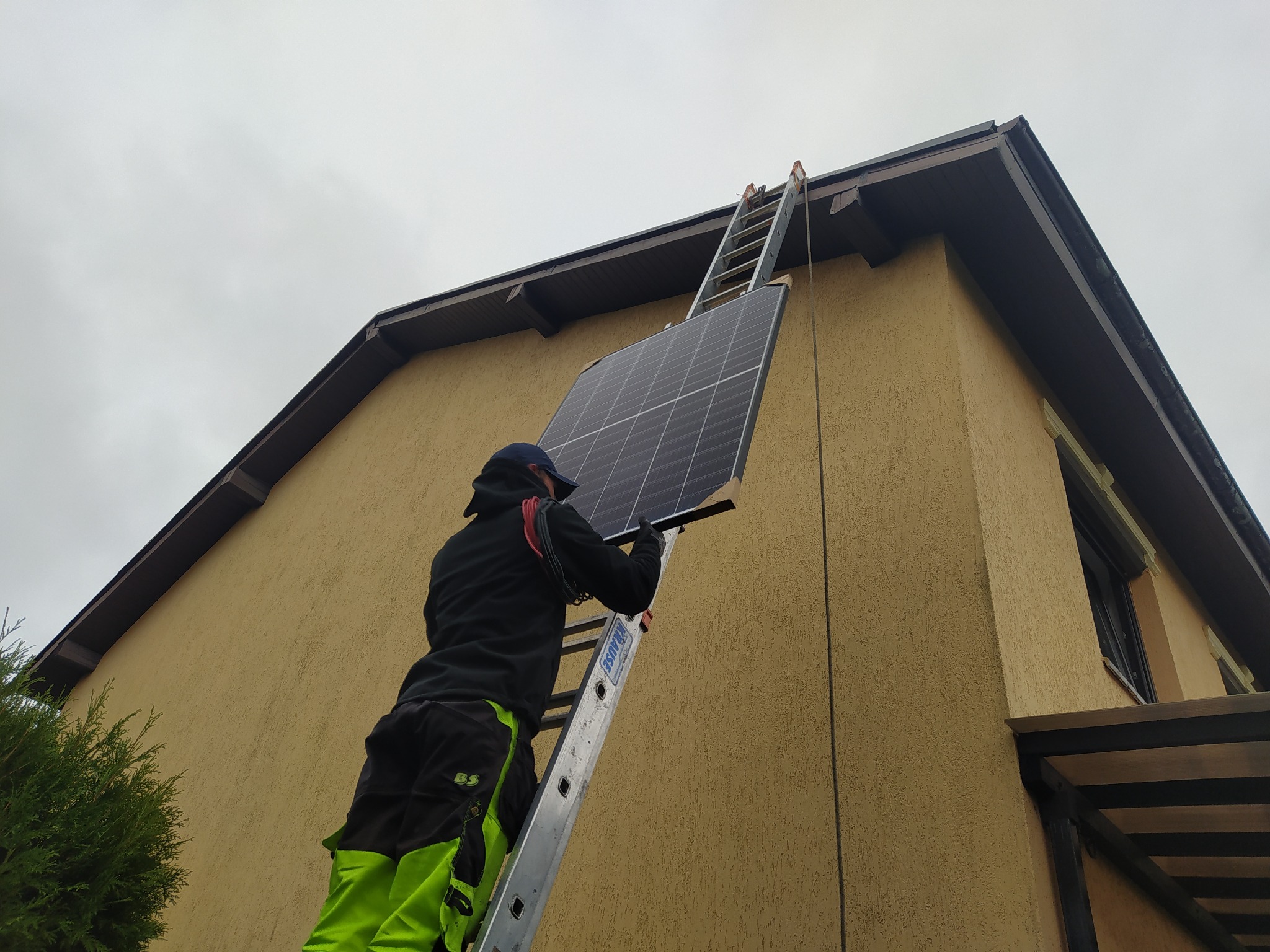 Instalator na drabinie przymocowuje panel słoneczny do ściany budynku z żółtą elewacją i brązowym dachem przy pochmurnym niebie.