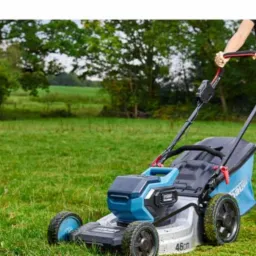 Elektryczna kosiarka do trawy marki Bauer na trawniku, obsługiwana przez osobę w ciemnych spodniach i koszulce, w tle drzewa i pole.