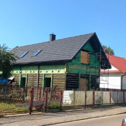 Dom w trakcie remontu elewacji, z czarnymi belkami i zielonymi deskami, nowy szary dach z oknami dachowymi, widoczne rusztowanie przy ścianie bocznej.