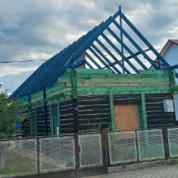 Konstrukcja drewnianego domu z widoczną więźbą dachową w kolorze niebieskim, czarne ściany obite papą, zielona impregnacja drewna, ogrodzenie z siatki w tle pochmurne niebo.