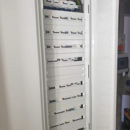 Krzysztof Bladowski - Otwarta, biała szafa elektryczna z wieloma zabezpieczeniami nadprądowymi i różnicowoprądowymi, widoczne okablowanie w górnej części, obok ściana w stanie surowym.