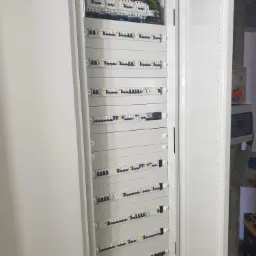 Otwarta, biała szafa elektryczna z wieloma zabezpieczeniami nadprądowymi i różnicowoprądowymi, widoczne okablowanie w górnej części, obok ściana w stanie surowym.