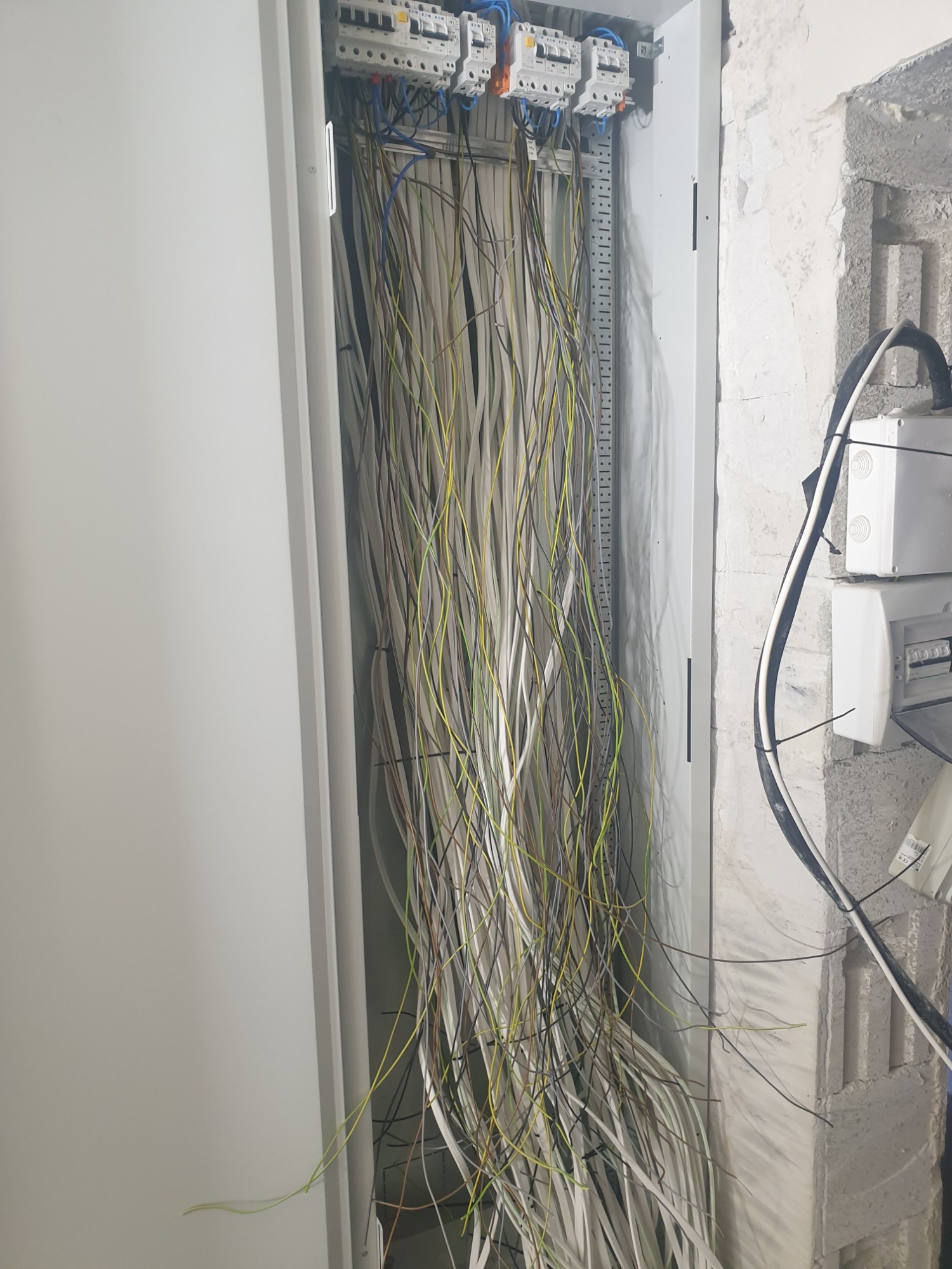 Otwarta szafa rozdzielcza z widocznymi przewodami elektrycznymi w różnych kolorach, zamontowana na surowej ścianie z betonu komórkowego.