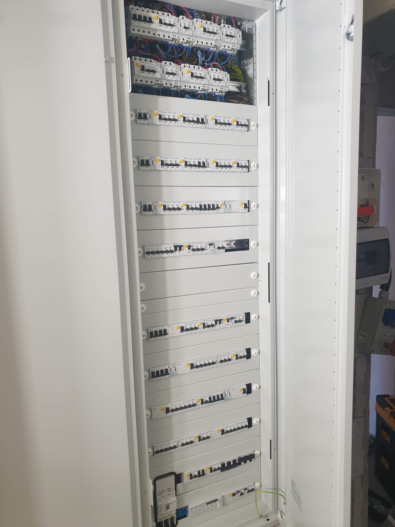 Otwarta, biała szafa elektryczna z wieloma zabezpieczeniami nadprądowymi i różnicowoprądowymi, widoczne okablowanie w górnej części, obok ściana w stanie surowym.
