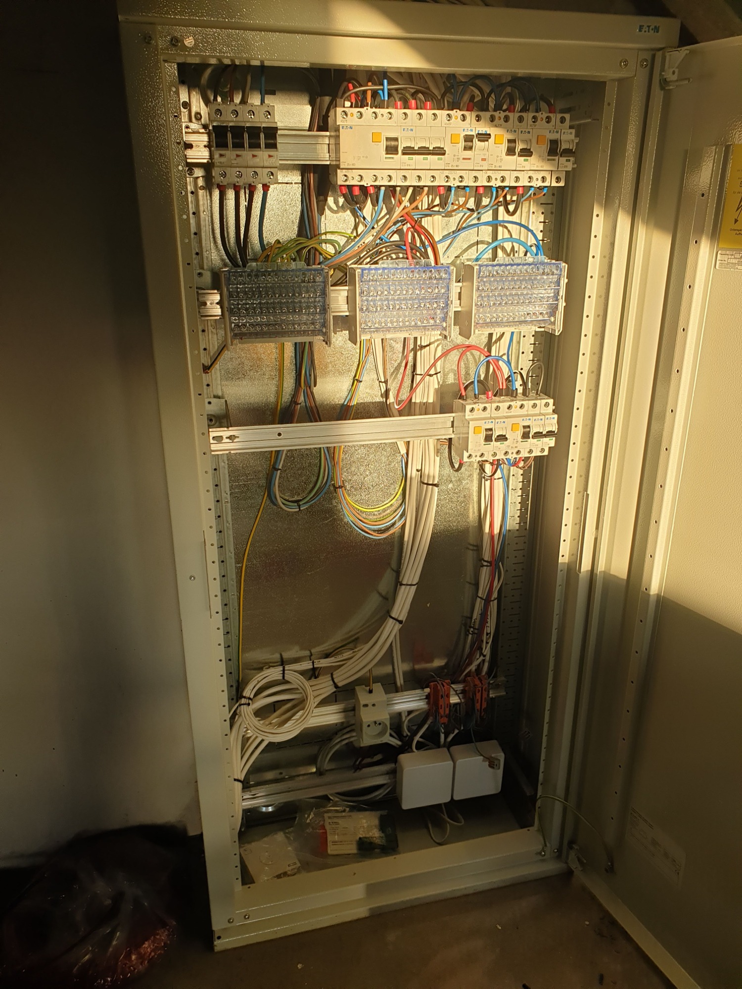 Otwarta szafa elektryczna z widocznymi zabezpieczeniami, przewodami różnej grubości i listwami zaciskowymi, oświetlona z góry.