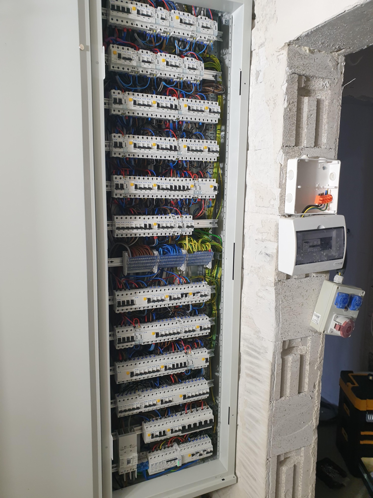 Rozdzielnica elektryczna z licznymi zabezpieczeniami nadprądowymi i różnicowoprądowymi, widoczne okablowanie, obok puszki instalacyjne na surowej ścianie z betonu komórkowego.