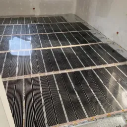 Rozłożone maty grzewcze na podłodze w surowym stanie, przygotowane do dalszych prac wykończeniowych, widoczne kable elektryczne i białe ściany.