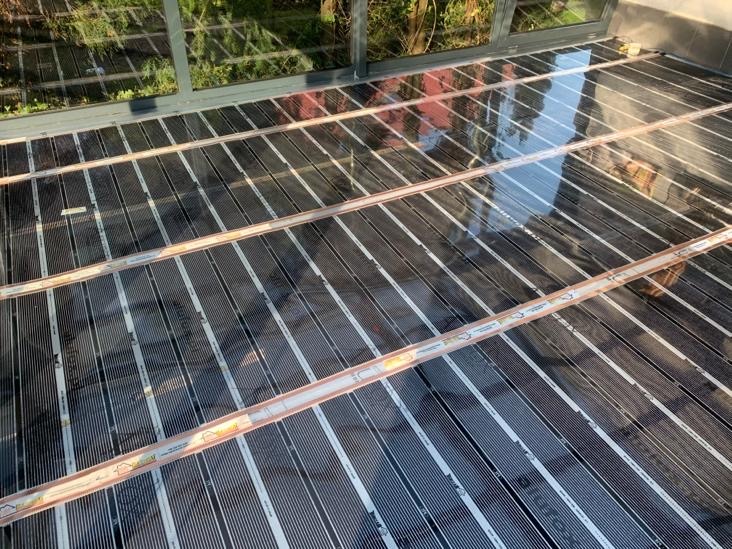 Rozłożone maty grzewcze na podłodze w pomieszczeniu z dużym oknem, widoczne elementy mocujące i odbicie otoczenia w powierzchni mat.