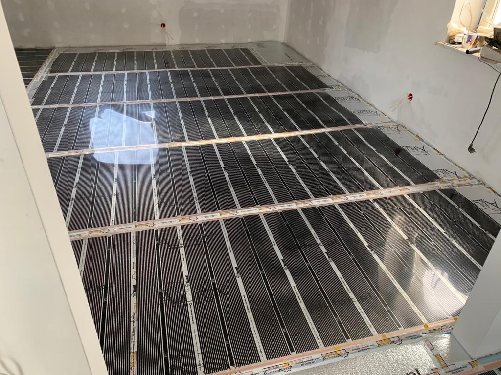 Rozłożone maty grzewcze na podłodze w surowym stanie, przygotowane do dalszych prac wykończeniowych, widoczne kable elektryczne i białe ściany.