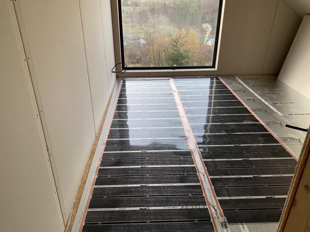 Montaż elektrycznego ogrzewania podłogowego: czarne maty grzewcze rozłożone na przygotowanym podłożu w pomieszczeniu z oknem, widoczne kable i folia aluminiowa.