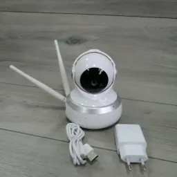 Biała kamera IP z dwiema antenami, kablem USB i zasilaczem, leżąca na szarym tle imitującym drewno.