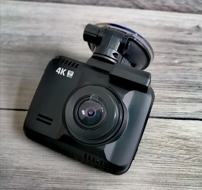 Czarna kamera samochodowa 4K WiFi z przyssawką na tle szarego drewnianego blatu, widok z góry, obiektyw z folią ochronną.