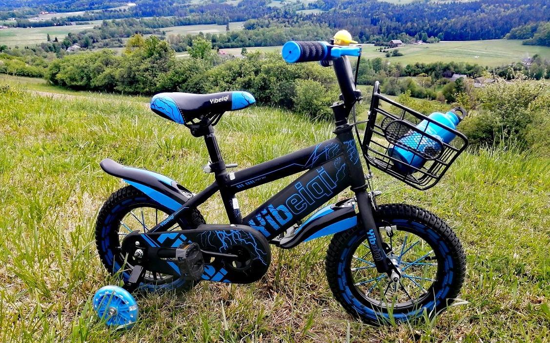 Czarno-niebieski rower dziecięcy z bidonem w koszyku, ustawiony na trawie z widokiem na pagórkowaty krajobraz w tle.