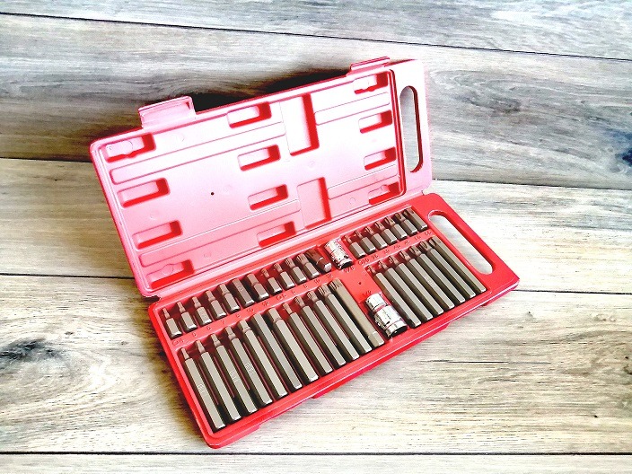 Otwarty, czerwony organizer z kompletem bitów i nasadek na tle szarej, drewnianej powierzchni.