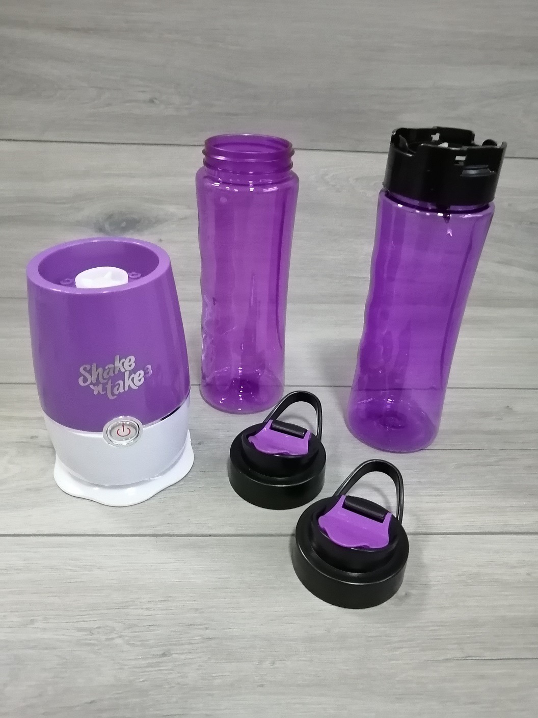 Fioletowy blender Shake'n'Take 3 z dwoma butelkami i zakrętkami na szarym tle drewnopodobnym.
