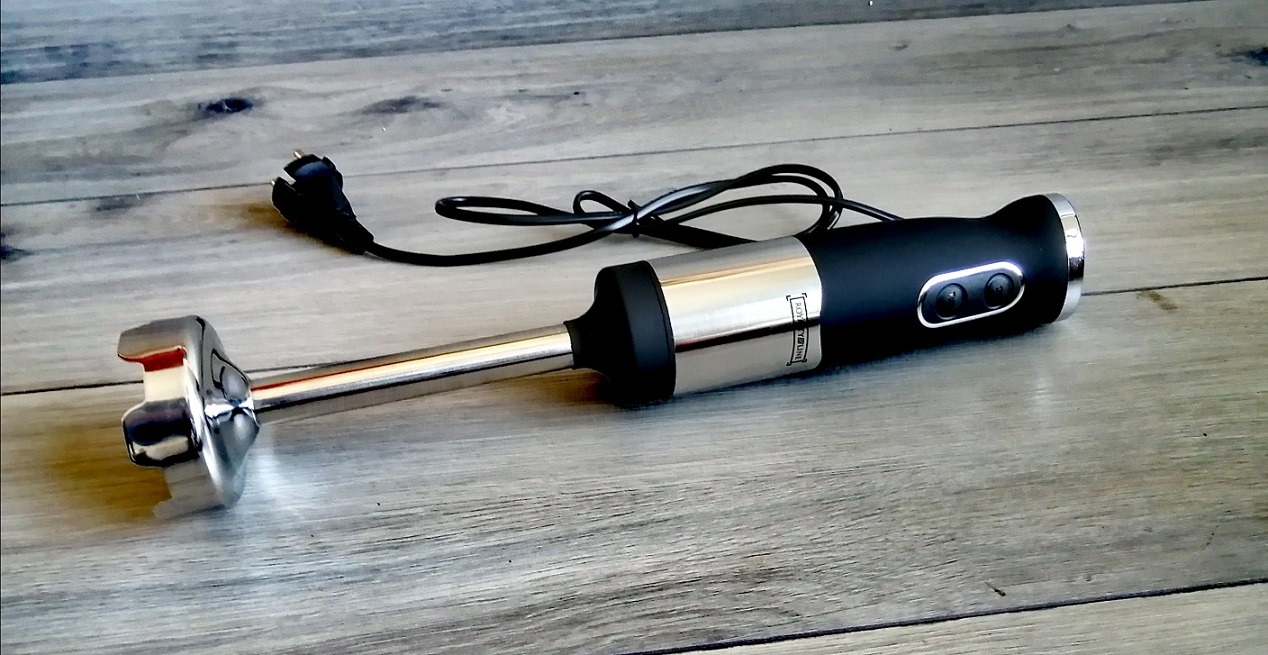 Czarny blender ręczny z metalową końcówką, leżący na szarym drewnopodobnym blacie, z widoczną wtyczką kabla zasilającego.