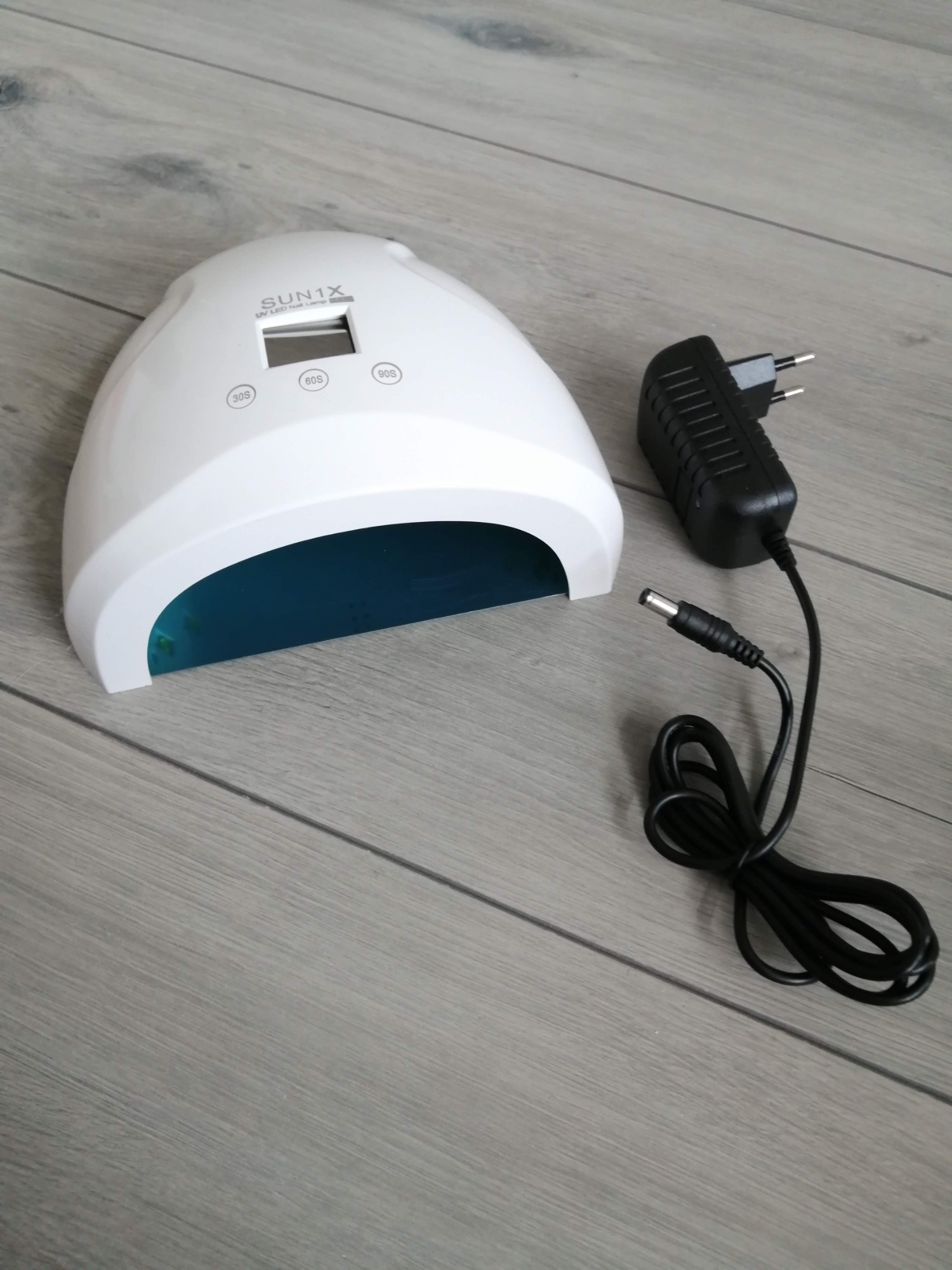 Biała lampa UV LED do paznokci marki SUN1X z wyświetlaczem i zasilaczem na szarym panelu podłogowym.