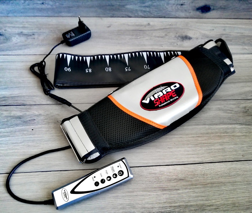 Pas wibrujący Vibro Shape z pilotem i miarką, leżący na drewnianym tle.