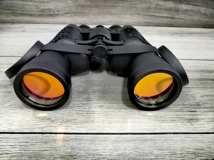 Czarne lornetki z pomarańczowymi soczewkami na szarym, drewnianym tle, widoczne z przodu.