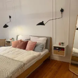 Nowoczesna sypialnia z drewnianą podłogą, łóżkiem z szarą ramą i pastelowymi poduszkami, oświetlona czarnymi lampami ściennymi o regulowanych ramionach, z lustrem stojącym w drewnianej ramie...