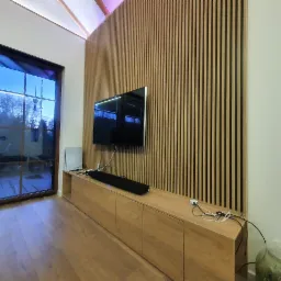Ściana z drewnianych lameli za telewizorem i szafką RTV z drewnianymi frontami, widok na okno z widokiem na zewnątrz.