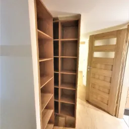 Wysoka, drewniana półka wnękowa z licznymi przegródkami, zamontowana w przedpokoju, z widocznymi drewnianymi drzwiami z przeszkleniami w tle.