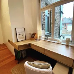 Drewniany blat biurka na wymiar, wkomponowany w przestrzeń przy oknie, z szufladami i półkami, uzupełniony o krzesło z poduszką i dekoracje okienne.