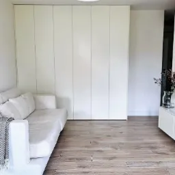 Jasne wnętrze pokoju dziennego z dużą, białą szafą w zabudowie, drewnianą podłogą i sofą z narzutą w geometryczne wzory.