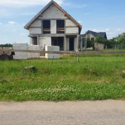 Surowy stan domu jednorodzinnego w trakcie budowy, widoczne bloczki betonowe, niezadaszona konstrukcja, drewniane elementy więźby dachowej, trawa i fragment drogi na pierwszym planie.